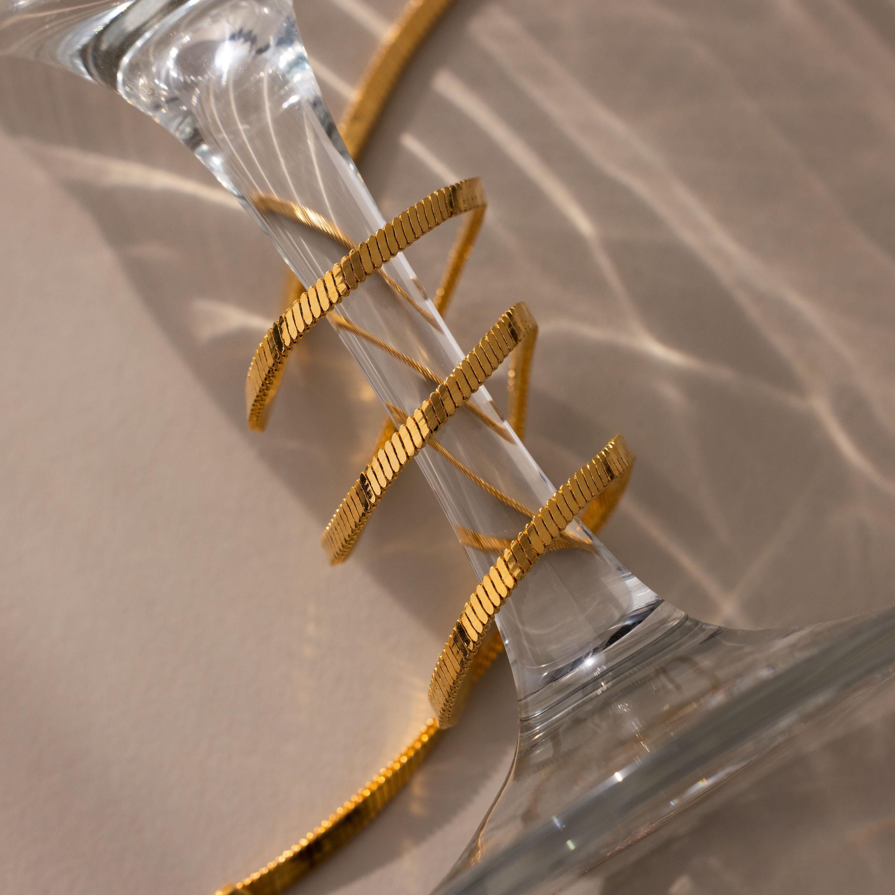 The Bold Snake Chain Necklace, a modern jewelry piece, is elegantly draped around the stem of an upside-down glass on a light surface.