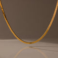 The Bold Snake Chain Necklace in 18K Gold is showcased on a neutral brown background with a soft shadow beneath, highlighting its modern design.