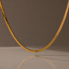 The Bold Snake Chain Necklace in 18K Gold is showcased on a neutral brown background with a soft shadow beneath, highlighting its modern design.