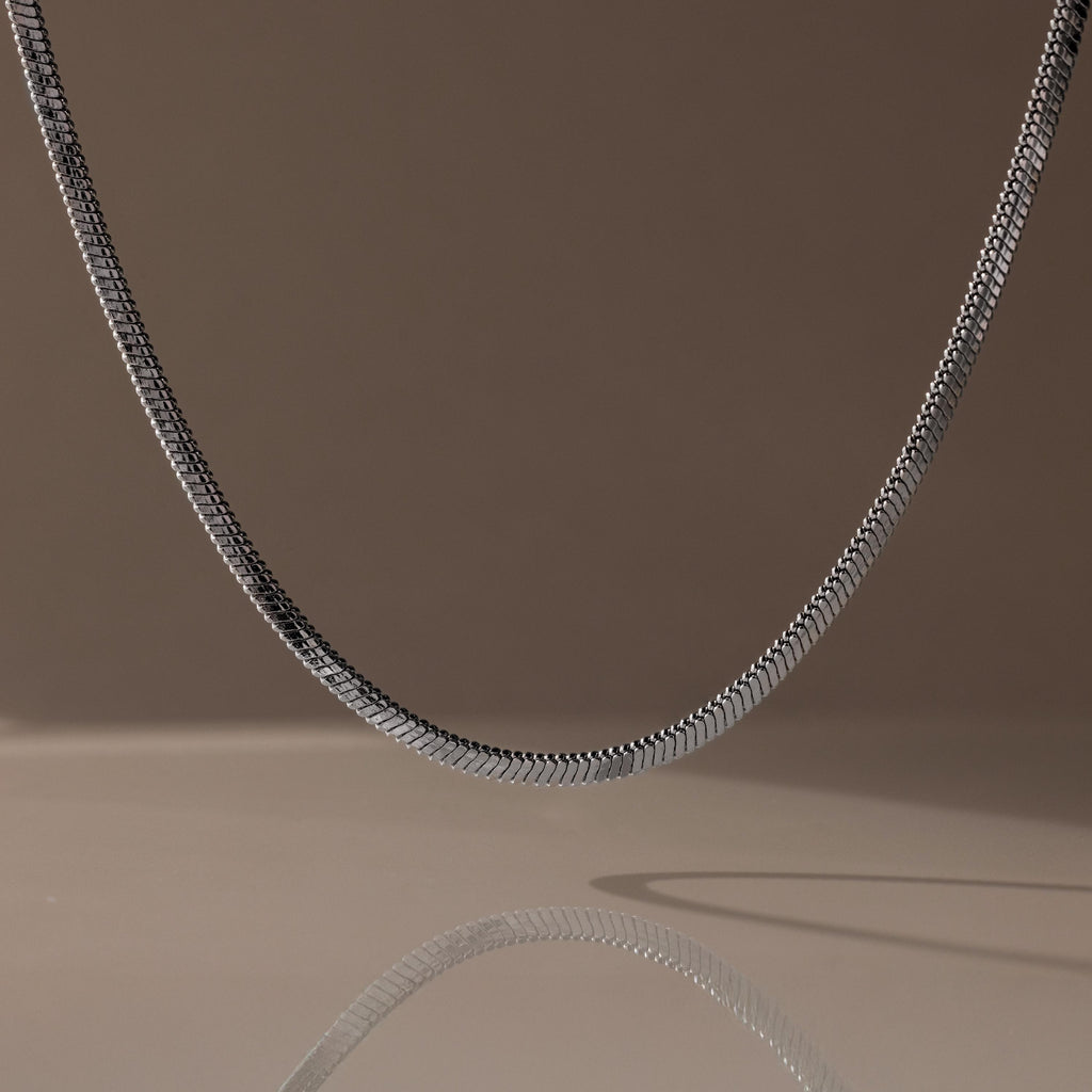 The Twisted Snake Chain Necklace in Silver hangs gracefully, its reflection shimmering on a smooth surface—an ideal choice for fans of modern jewelry.