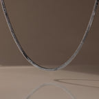 The Twisted Snake Chain Necklace in Silver hangs gracefully, its reflection shimmering on a smooth surface—an ideal choice for fans of modern jewelry.
