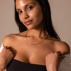Woman with long dark hair smiles softly, wearing a black top, white jacket off her shoulders, and the Bold Snake Chain Necklace in 18K Gold.