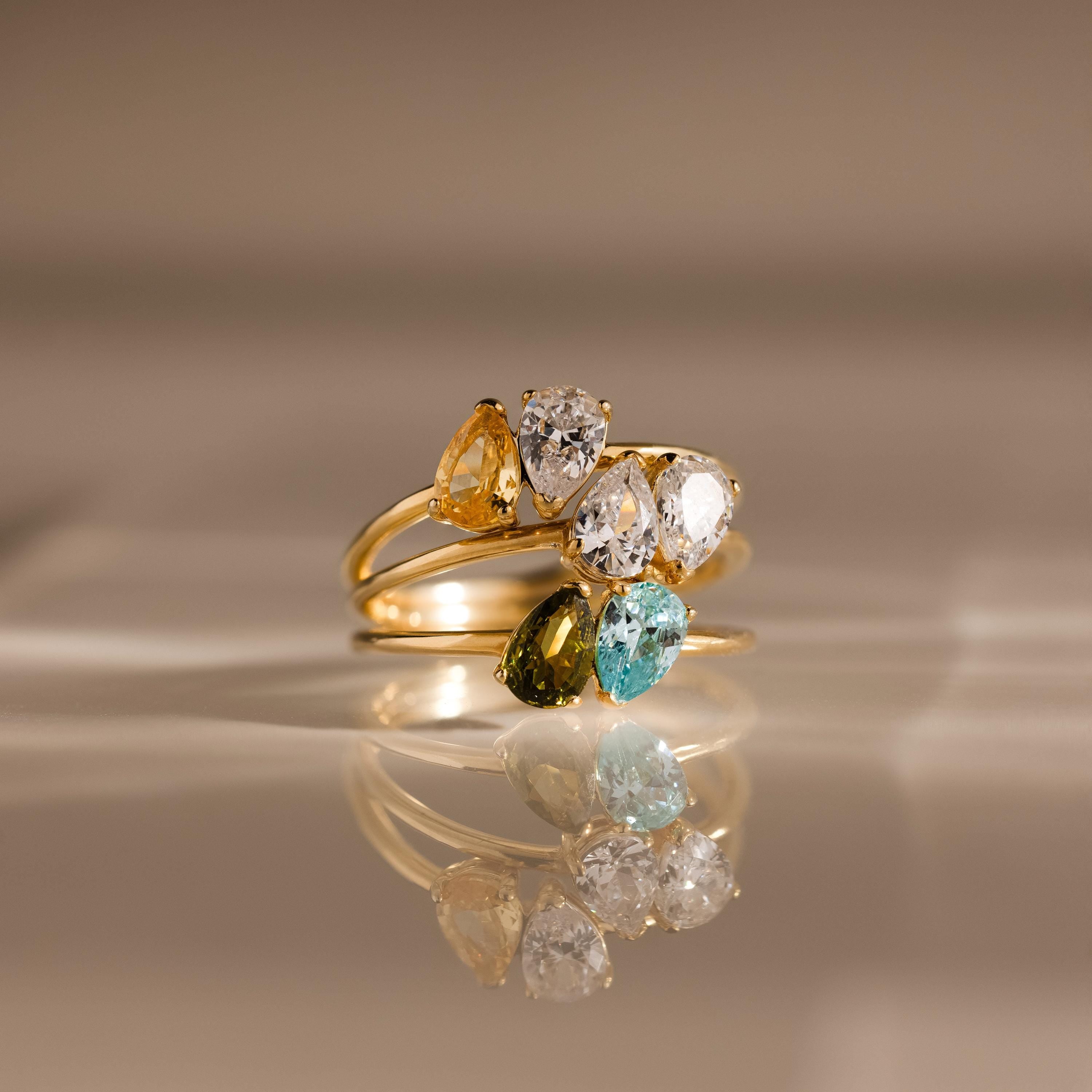 Two 18K gold Duo Pear Toi Et Moi Rings with pear-shaped stones in yellow, clear, green, and blue are displayed on a reflective surface, showcasing their romantic and enchanting appeal.