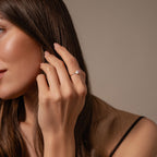 A woman with long brown hair touches her face with her left hand, wearing the Duo Pear Toi Et Moi Ring in Silver adorned with pear-shaped stones.