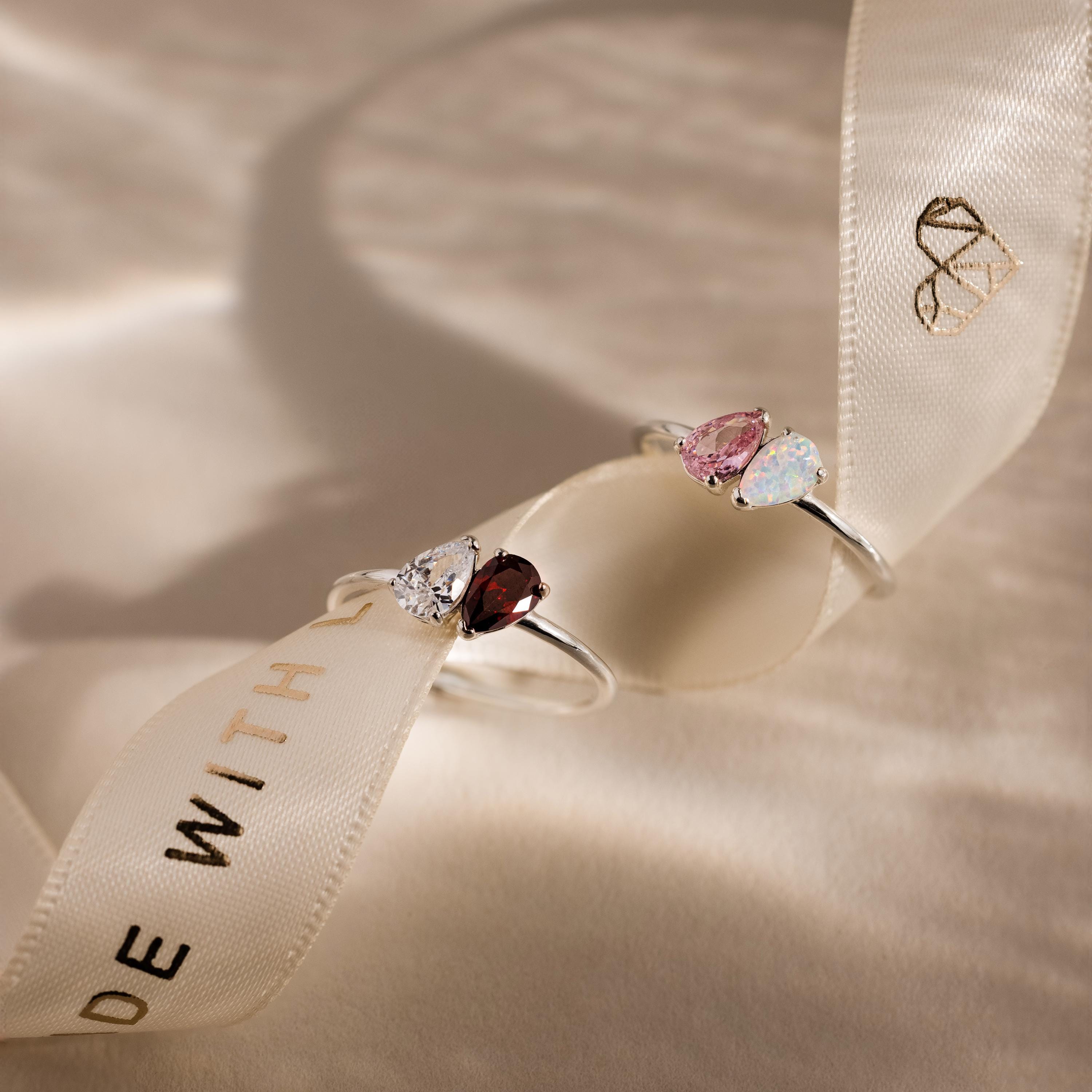 Two Duo Pear Toi Et Moi Rings in Silver, featuring colorful pear-shaped stones, are placed on a cream ribbon with gold lettering and a heart logo, creating a romantic display.