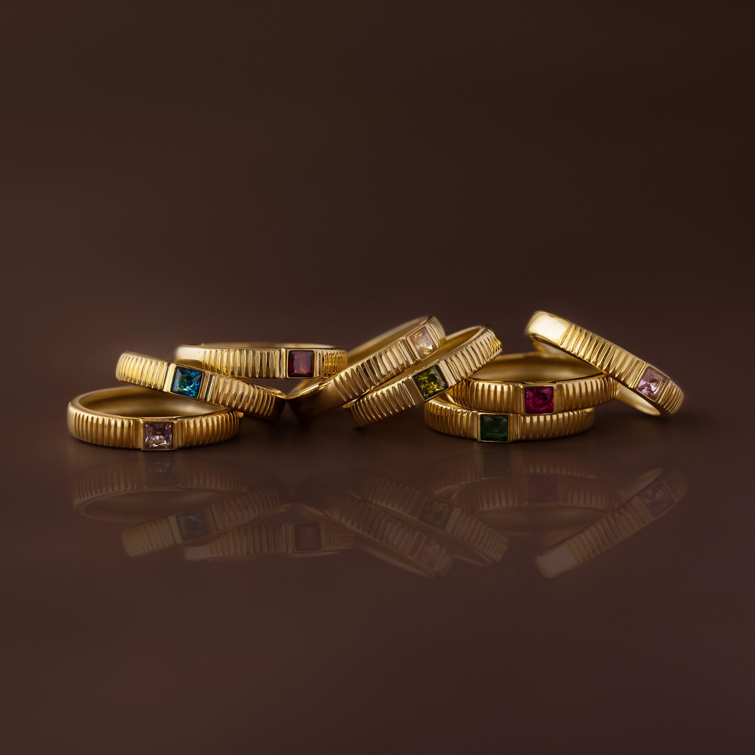 The Ribbed Opal Band is displayed on a brown background alongside other stacking rings with colorful gemstones, arranged in a semicircle.