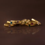 Eight gold stacking rings, one of which is the Ribbed Blue Zircon Band, along with birthstone rings featuring colored gemstones, are grouped together on a brown reflective surface.
