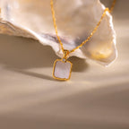 The Square Mother of Pearl Necklace features an iridescent pendant and a small diamond, showcased on a light surface next to a shell.