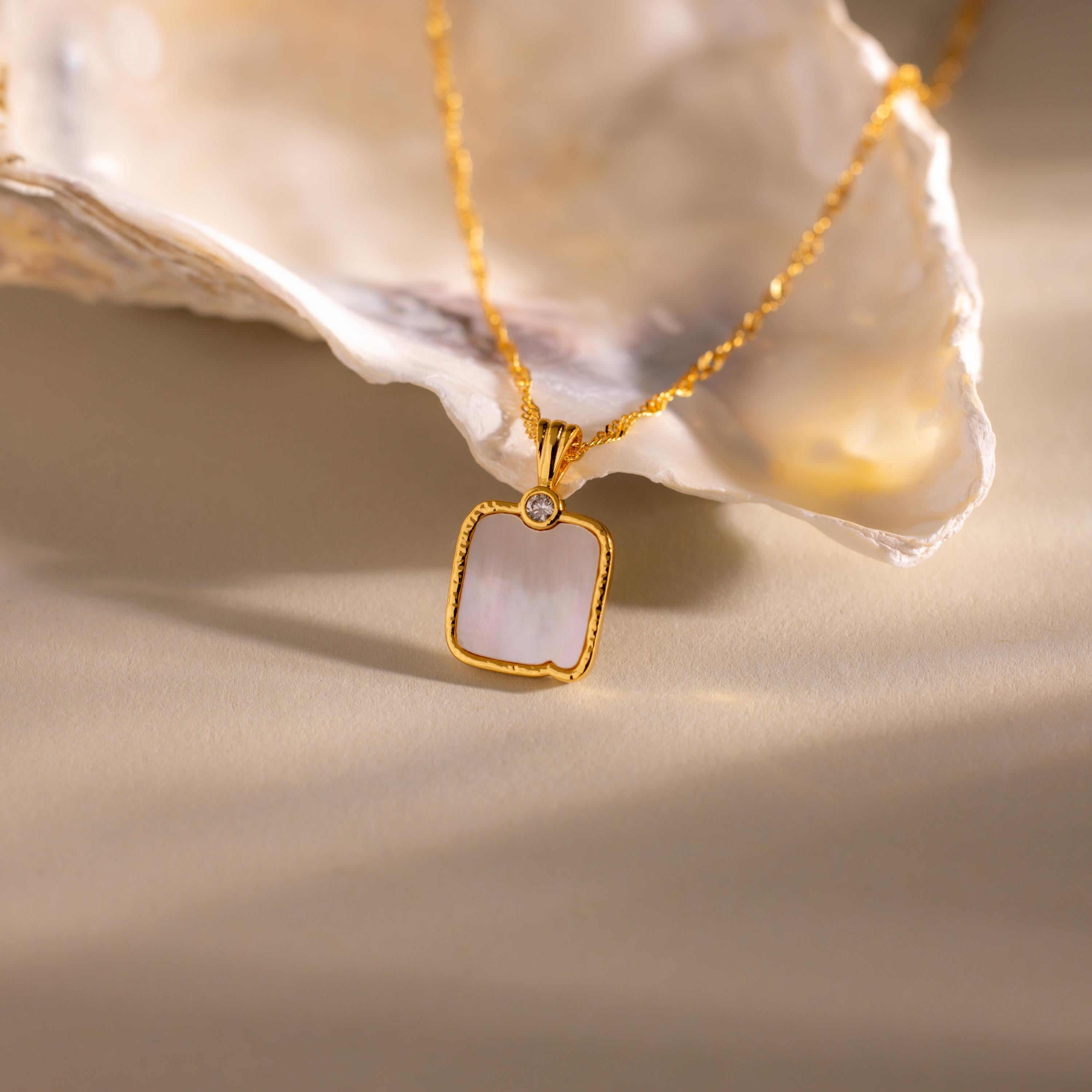The Square Mother of Pearl Necklace features an iridescent pendant and a small diamond, showcased on a light surface next to a shell.