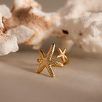 The Open Duo Starfish Ring sits on a beige surface with white coral behind it, capturing coastal charm and making a perfect statement piece.
