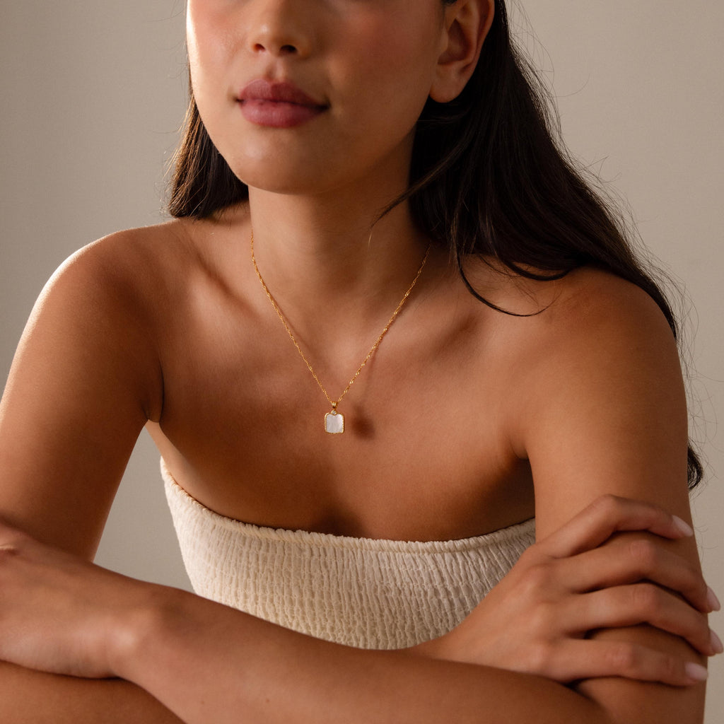 A woman in a strapless textured top wears the Square Mother of Pearl Necklace, a delicate gold jewelry piece, as she sits with her arms crossed.