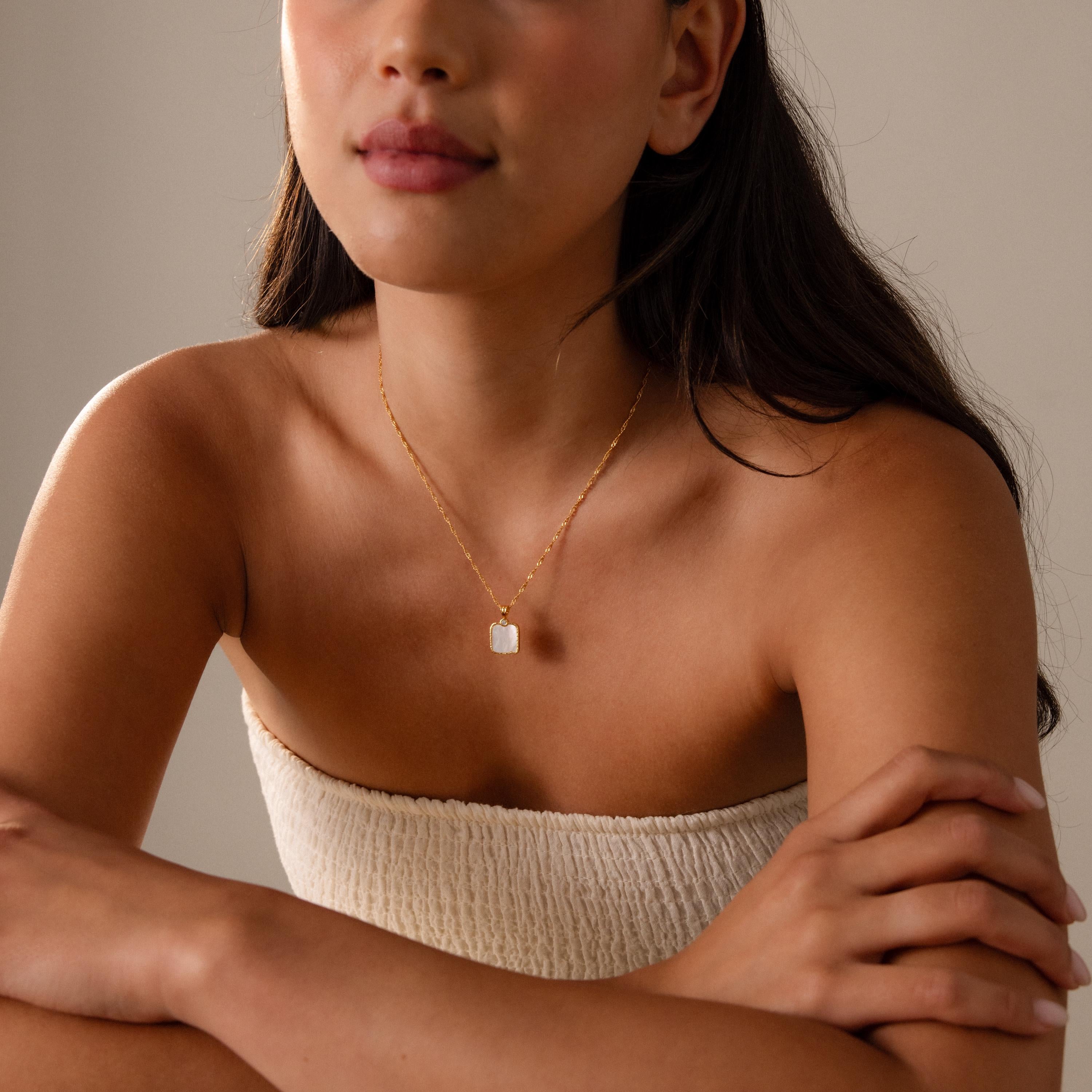 A woman in a strapless textured top wears the Square Mother of Pearl Necklace, a delicate gold jewelry piece, as she sits with her arms crossed.