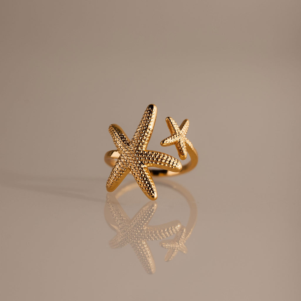 The Open Duo Starfish Ring showcases two textured starfish, one large and one small, on a reflective beige surface—a stunning gold coastal-inspired statement jewelry piece.