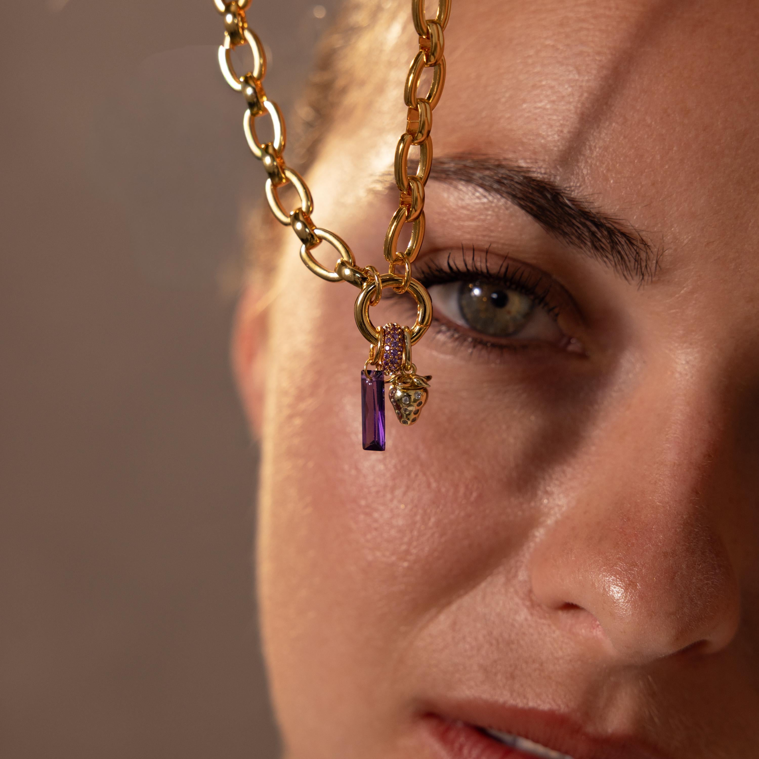 A gold chain necklace with purple and gold birthstone charms and a Pave Amethyst Charm Spacer is held near a womans face, highlighting her eye.