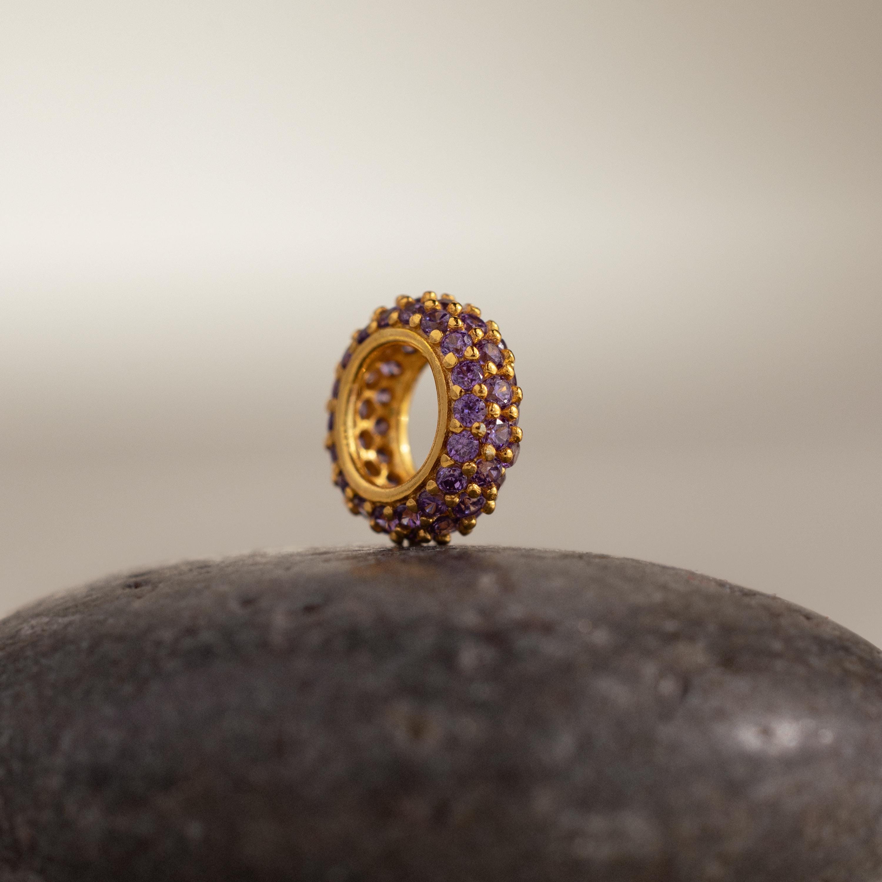 The Pave Amethyst Charm Spacer, adorned with purple stones, stands upright on a smooth, dark stone.