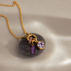 Gold necklace featuring a Hexagon Alexandrite Charm and purple gemstone accents, each set in geometric frames, displayed on a smooth gray stone against a neutral background.