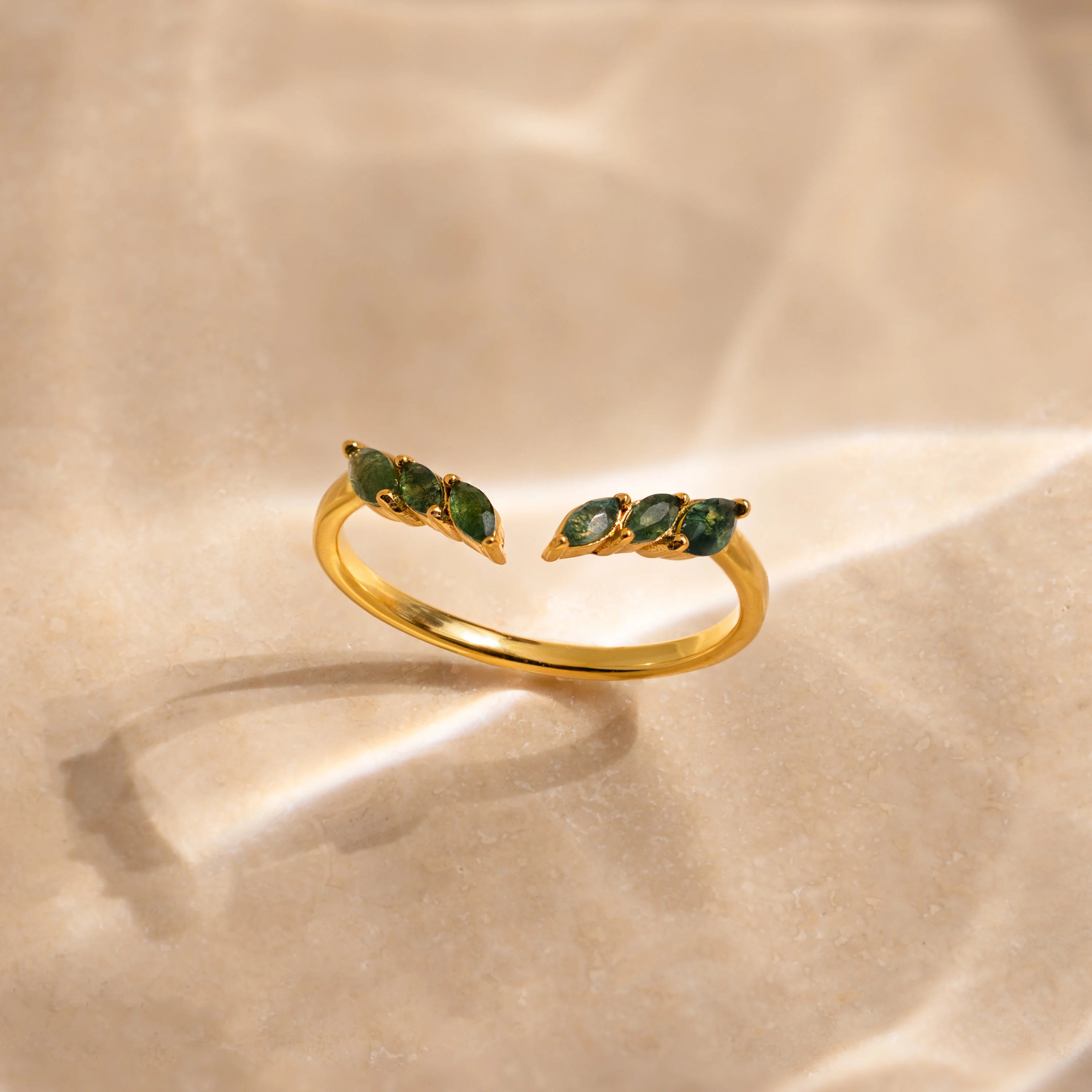 The Open Moss Agate Chevron Ring features green gemstone accents and is showcased on a beige sunlit surface with soft shadows—a perfect choice for a dainty ring or promise ring.