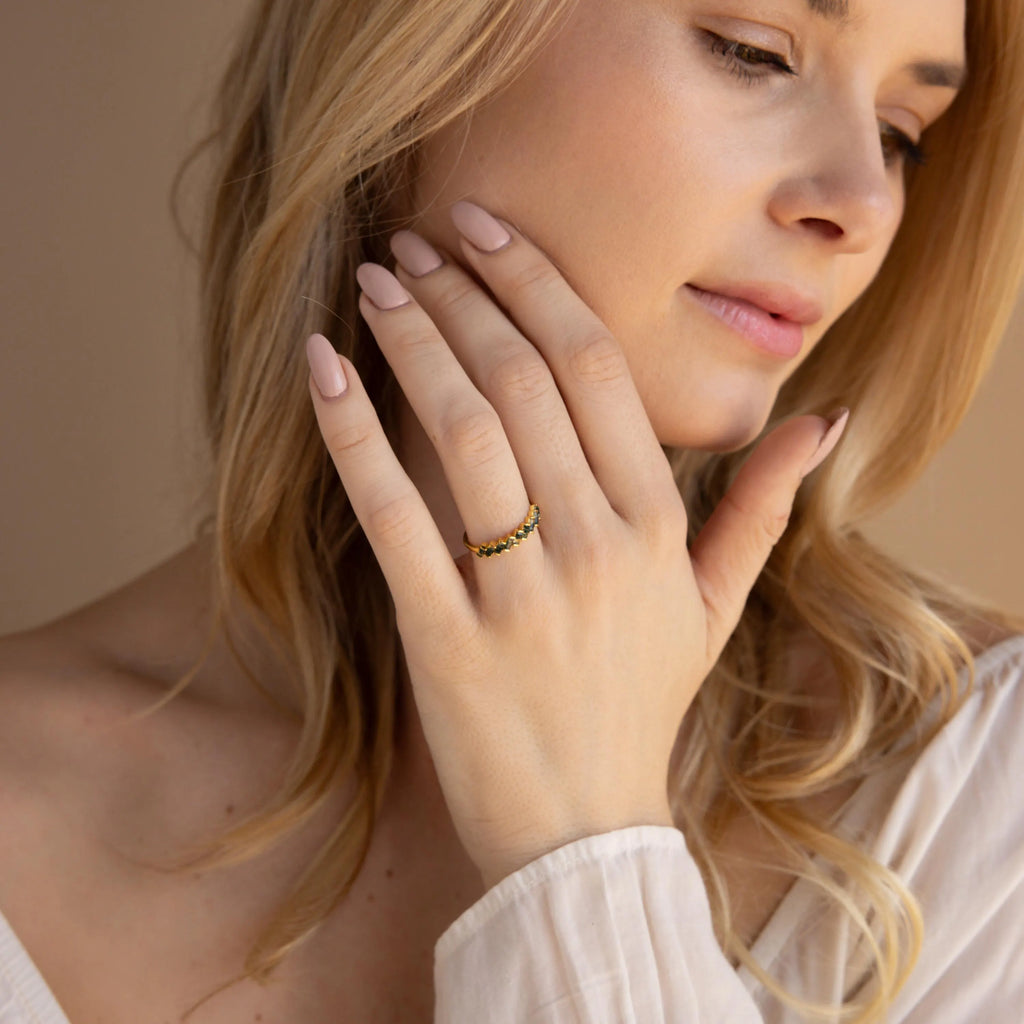 A woman with long blonde hair wears the Moss Agate Eternity Ring, gently touching her face and looking down in soft natural light.