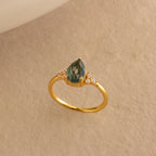 The Agate Teardrop Ring in 18K Gold features a pear-shaped green gemstone and small diamonds, set on a beige textured background—reminiscent of a vintage cocktail ring and perfect for making a statement.