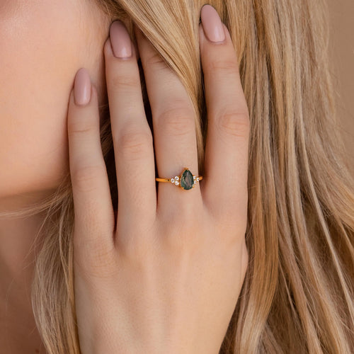 A hand with nude nails wears the Agate Teardrop Ring, showcasing a green moss agate gemstone and sparkling accent stones set in gold.