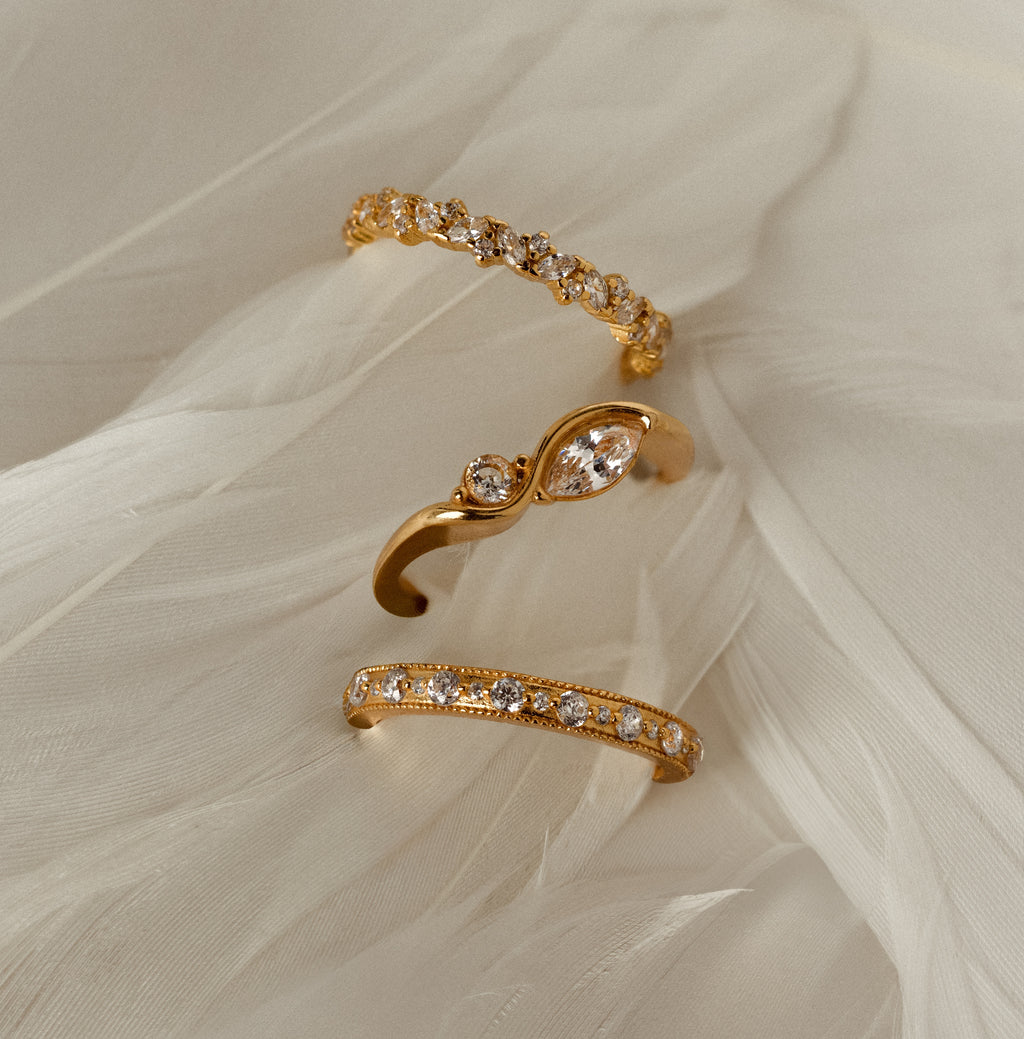 The Diamond Eternity Ring shines among three gold rings, set on soft white fabric with a feather-like texture.