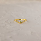 The Peridot Drop Curve Ring—a delicate gold stacking ring featuring a small green peridot gemstone—rests on a textured white surface.