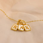 A gold chain necklace with three square pendant charms featuring white pearl inlay and raised gold letters 'H', 'A', and 'K', displayed on a soft beige fabric background.