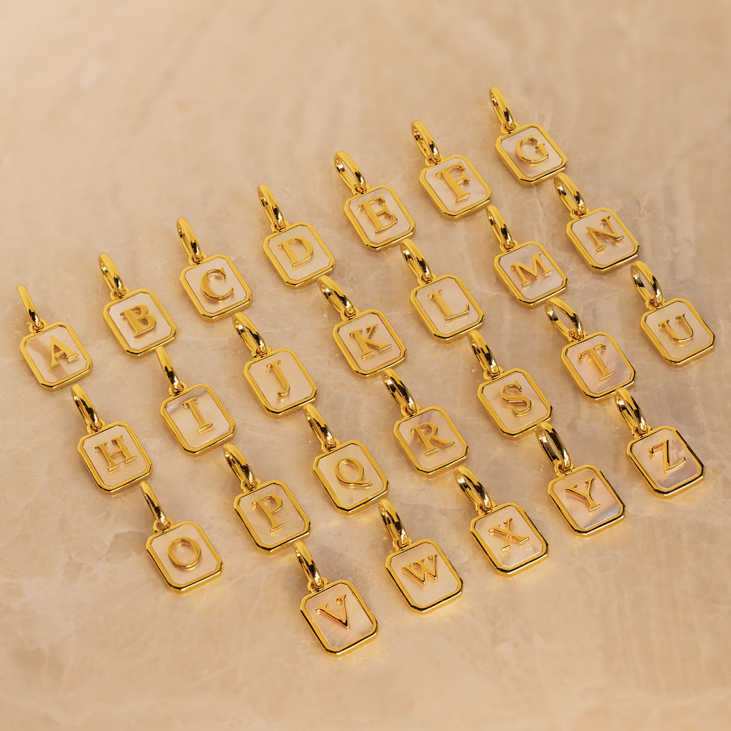 Gold-plated square letter charms spelling out the alphabet, each with a white inlay and a small lobster clasp at the top, arranged on a textured beige surface.