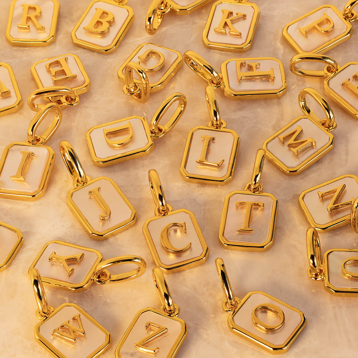 A close-up arrangement of gold letter charms with white pearl enamel backgrounds, each featuring a raised uppercase letter and a small jump ring, photographed on a light neutral surface.