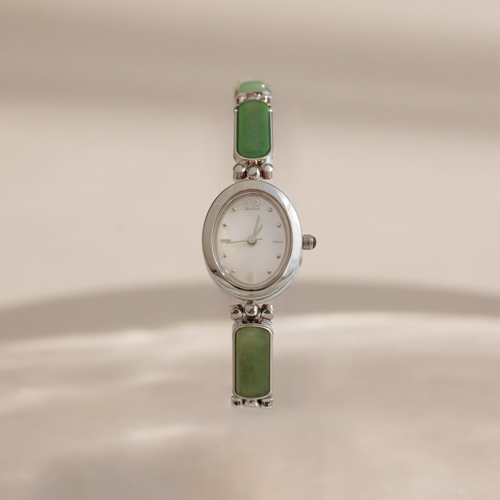 A vintage-style Jade Band Timepiece watch with a silver-tone oval case, white dial, and green rectangular stone links, shown against a warm beige background.