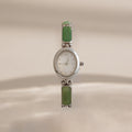 A vintage-style Jade Band Timepiece watch with a silver-tone oval case, white dial, and green rectangular stone links, shown against a warm beige background.