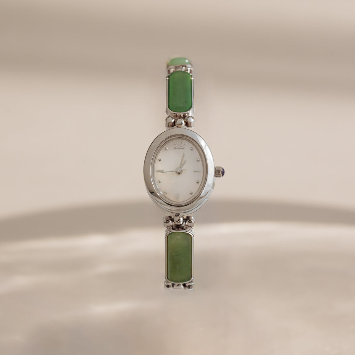 A vintage-style Jade Band Timepiece watch with a silver-tone oval case, white dial, and green rectangular stone links, shown against a warm beige background.