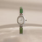A vintage-style Jade Band Timepiece watch with a silver-tone oval case, white dial, and green rectangular stone links on a matching bracelet, shown against a warm beige background.