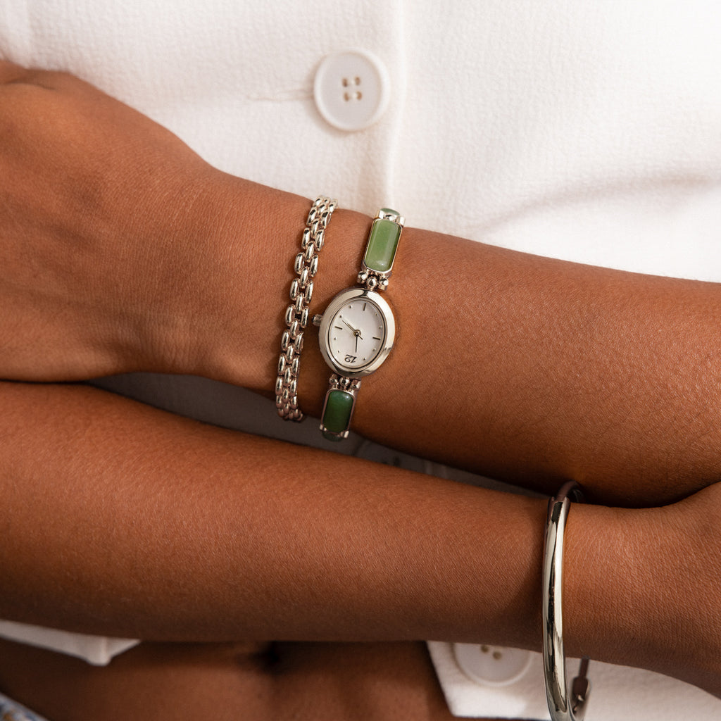 A close-up of a woman's wrist wearing our Jade Band Timepiece watch, layered over a silver chain bracelet and a thin silver bangle, against a white buttoned top.
