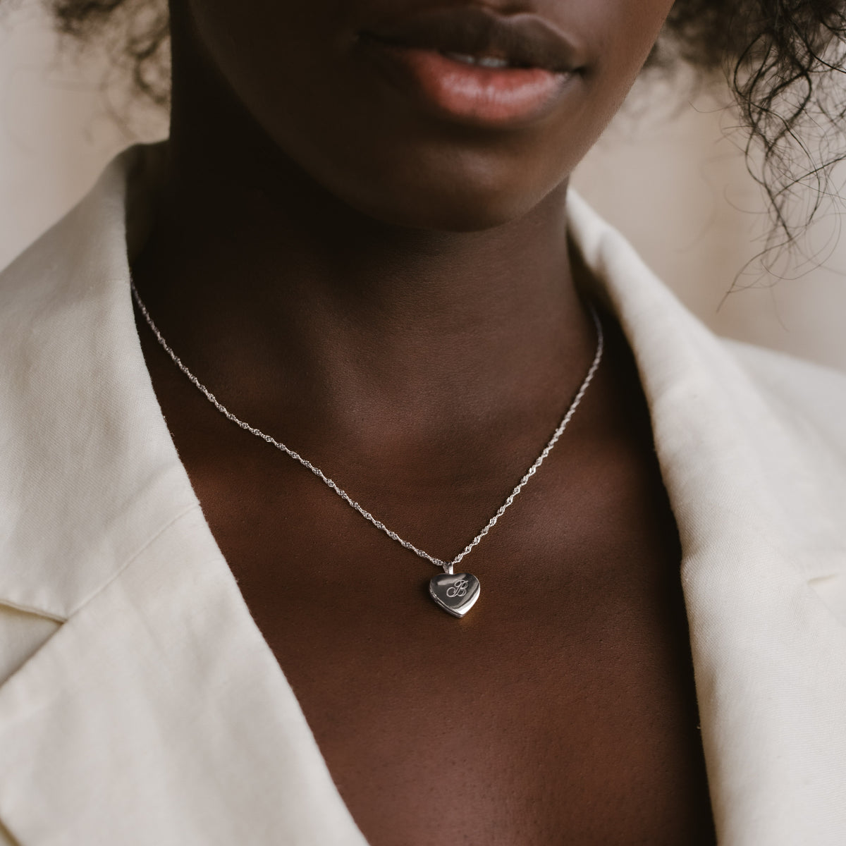 A close-up of a model wearing a delicate silver chain necklace with a small heart-shaped pendant engraved with the letter, styled with a white linen jacket.