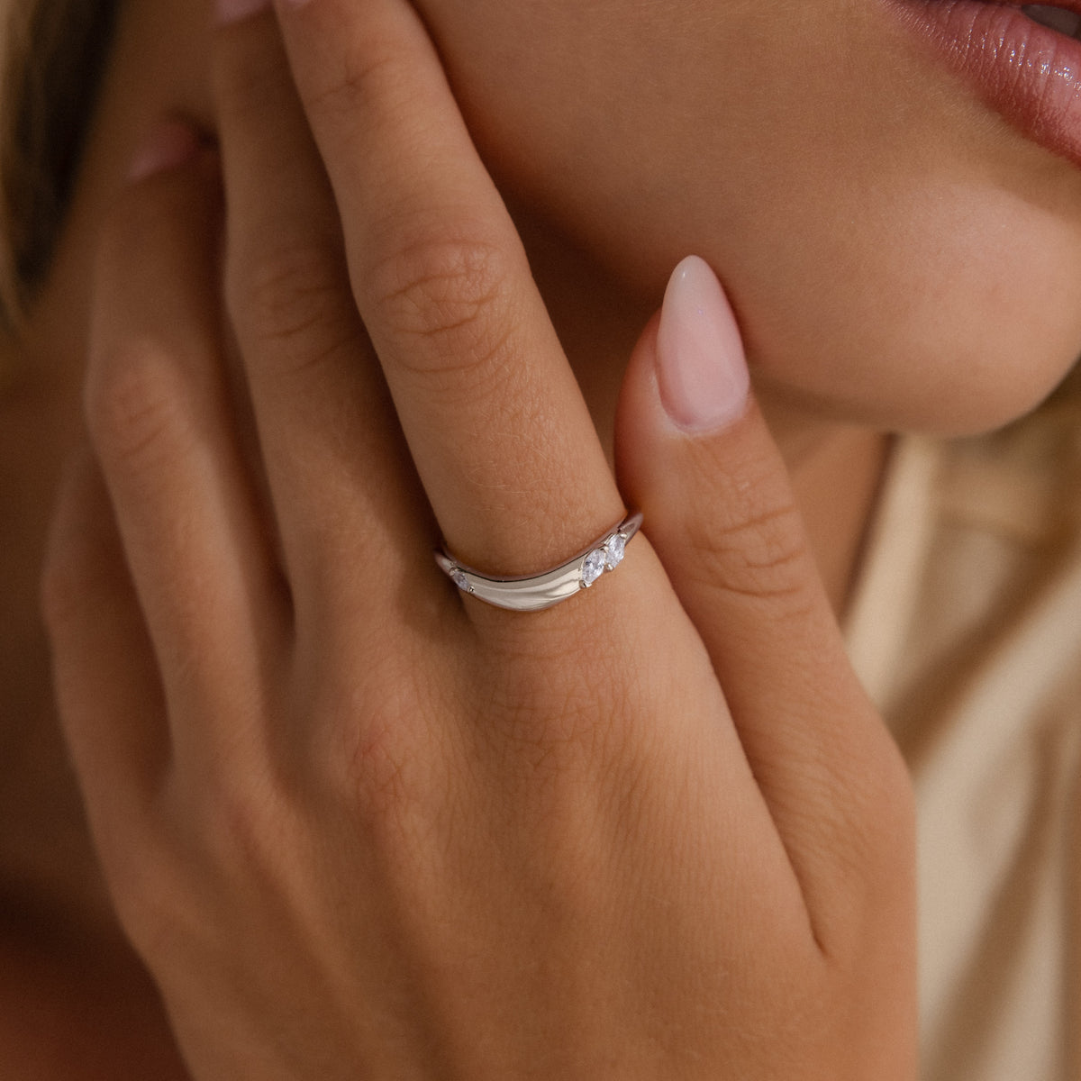 A close-up lifestyle shot of a woman's hand wearing a delicate silver band ring with a small clear gemstone, set against her face and shoulder.