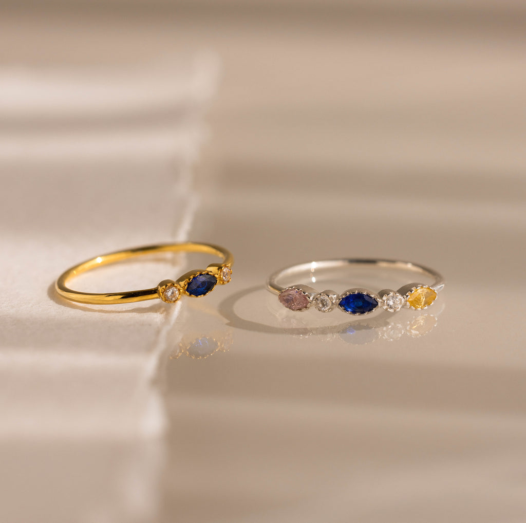Two slim rings on a soft beige surface: one gold ring with a deep blue oval stone and two small clear accent stones, and one silver ring with a blue oval stone, a small pink oval stone, and a small yellow oval stone with clear accent stones.
