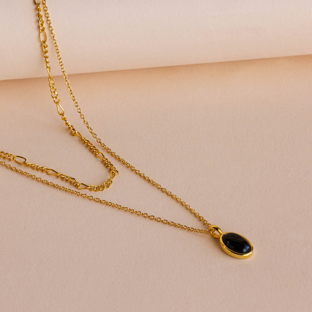 A gold layered necklace with a small oval black onyx pendant, displayed on a soft beige background.