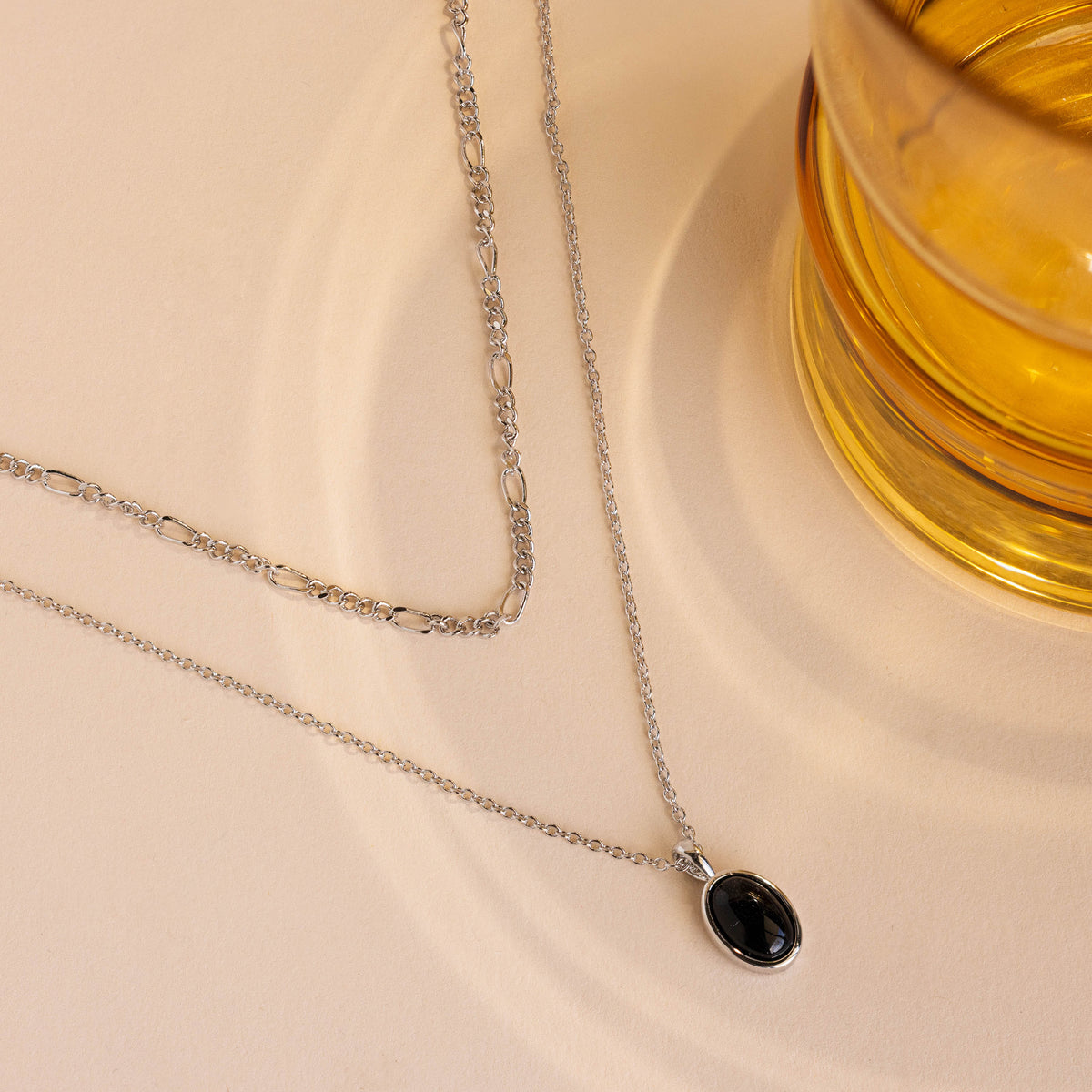A silver-toned double-layer necklace with a black oval pendant, styled on a beige textured surface next to a glass of amber-colored liquid.
