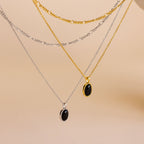 Three layered necklaces displayed on a neutral beige background: a silver chain with a black oval pendant, a gold chain with a black oval pendant, and a silver Figaro chain with a black oval pendant.