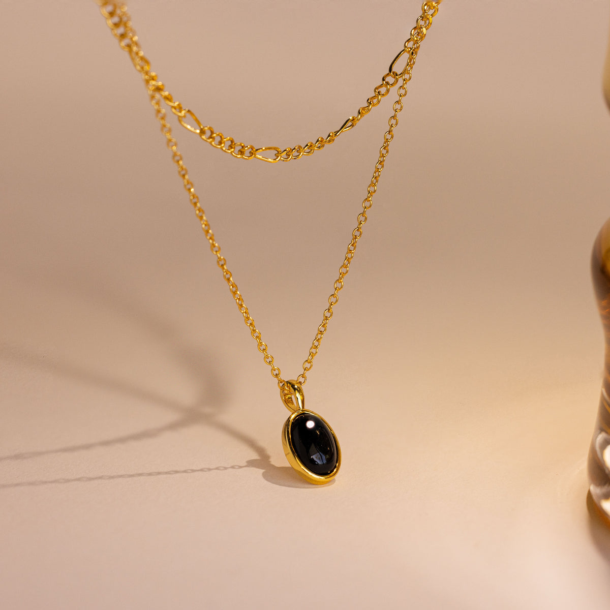 A gold layered necklace featuring a delicate chain and a longer chain with an oval black stone pendant, displayed on a warm beige surface.