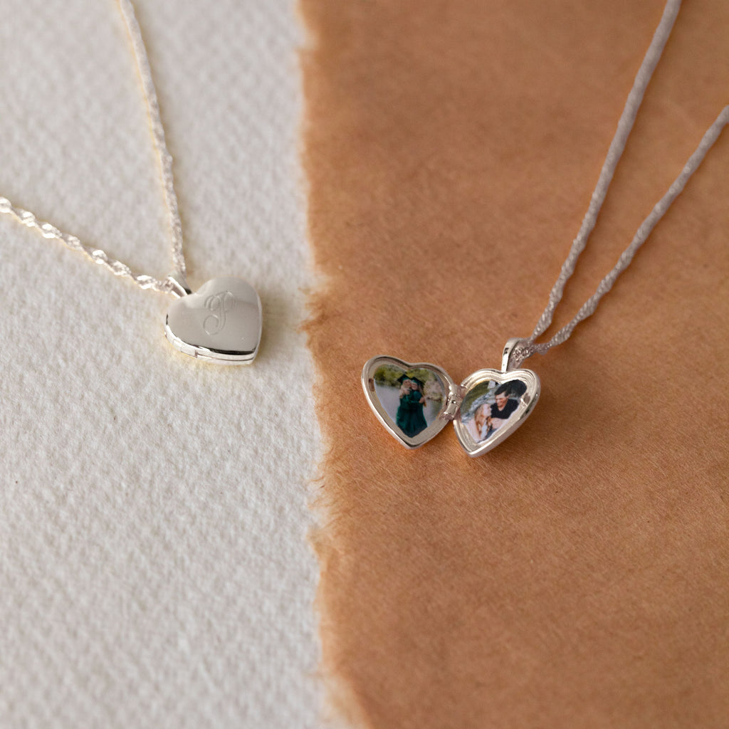 Two silver heart-shaped locket necklaces on delicate chains, displayed on a split white and warm brown textured background. One necklace features an engraved silver heart, while the other is open to reveal custom printed photos inside the heart.