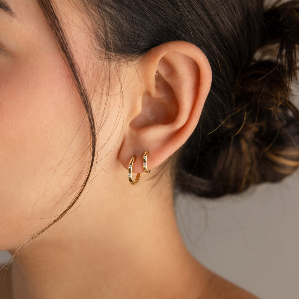 A close-up of a person's ear wearing a pair of small gold huggie hoop earrings set against a soft neutral background.