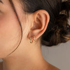 A close-up of a person's ear wearing a pair of small gold huggie hoop earrings set against a soft neutral background.