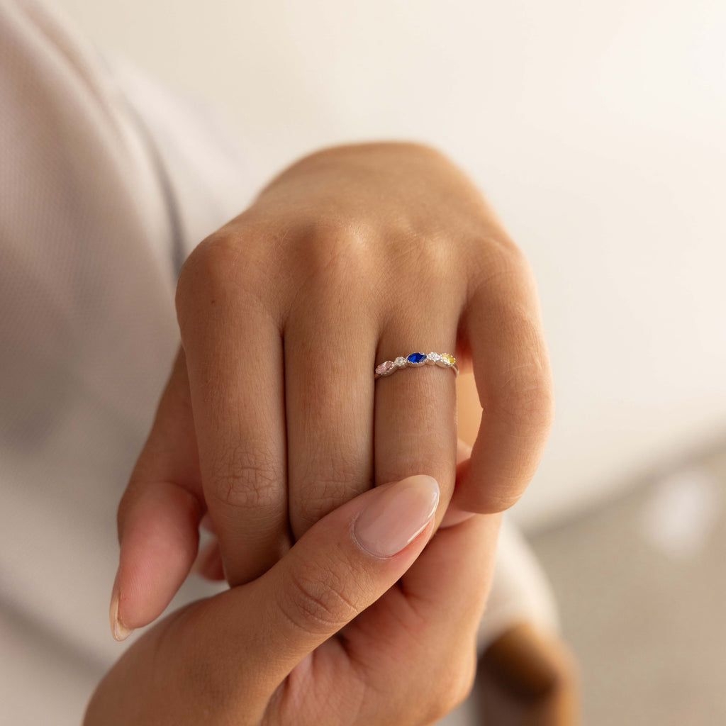 A close-up lifestyle shot of a delicate silver band ring worn on a person's ring finger. The ring features a small central blue oval gemstone flanked by tiny clear stones on each side.