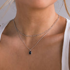A close-up of a model wearing a double-layered silver necklace. The top layer features a delicate curb chain, and the bottom layer features a fine link chain with an oval black pendant set in a silver bezel.