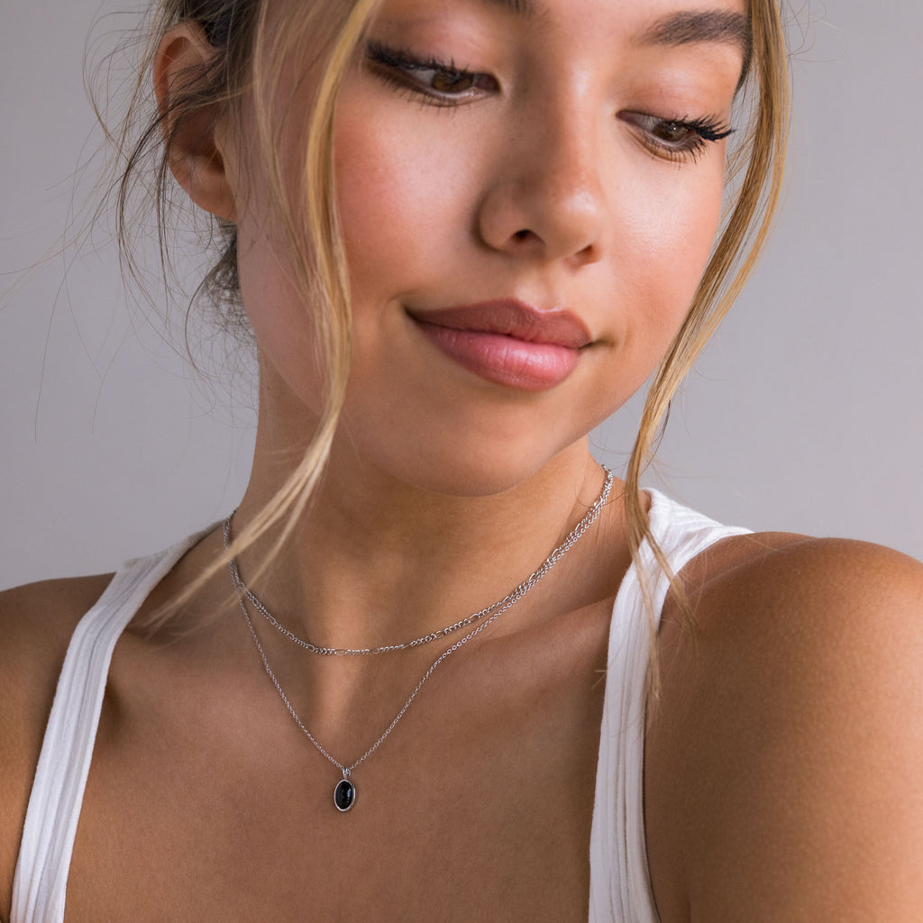 A close-up of a woman wearing a delicate silver double-layer necklace with a small oval dark blue stone pendant, paired with a white tank top against a neutral gray background.