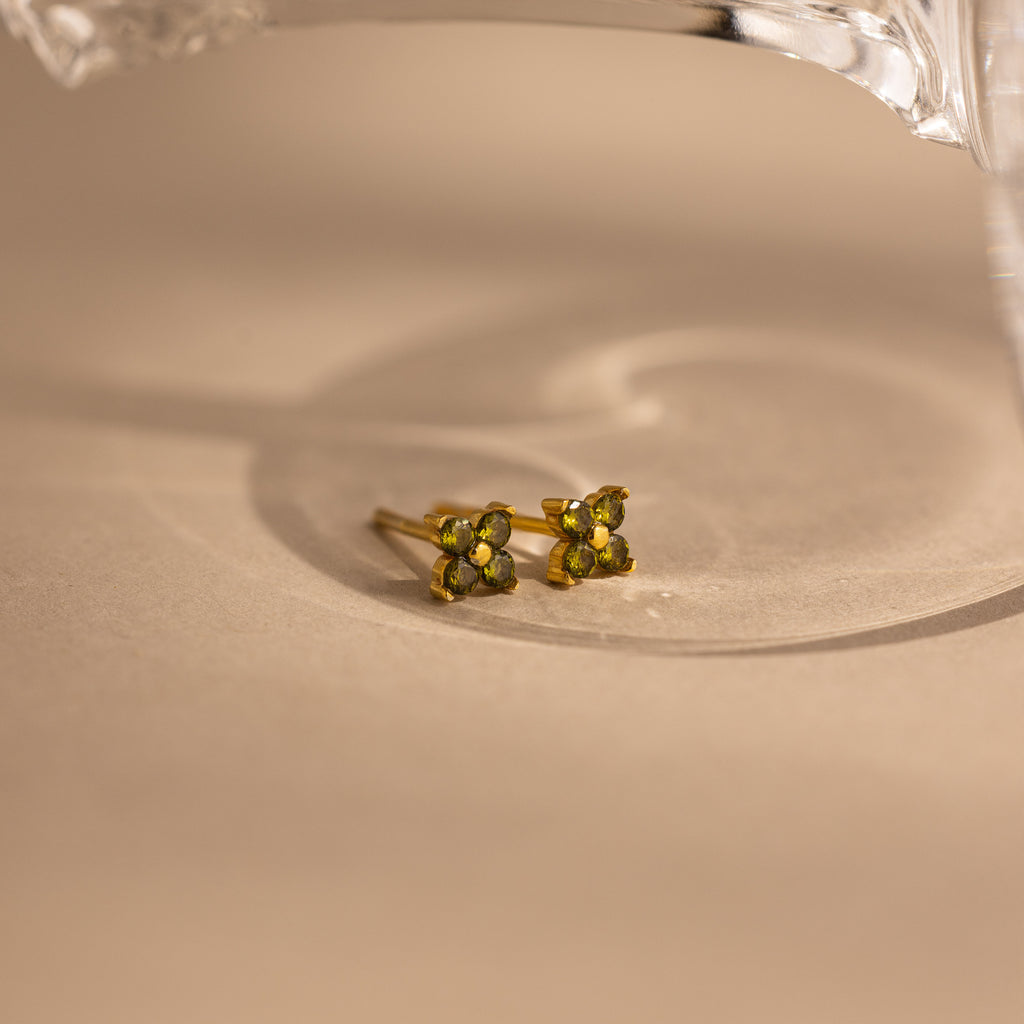 A pair of small gold stud earrings with four green gemstones arranged in a flower shape, resting on a beige surface with a soft shadow.