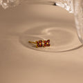 A pair of gold-toned stud earrings with four small red gemstones arranged in a square, resting on a beige surface with a soft shadow and a glass object partially visible at the top.