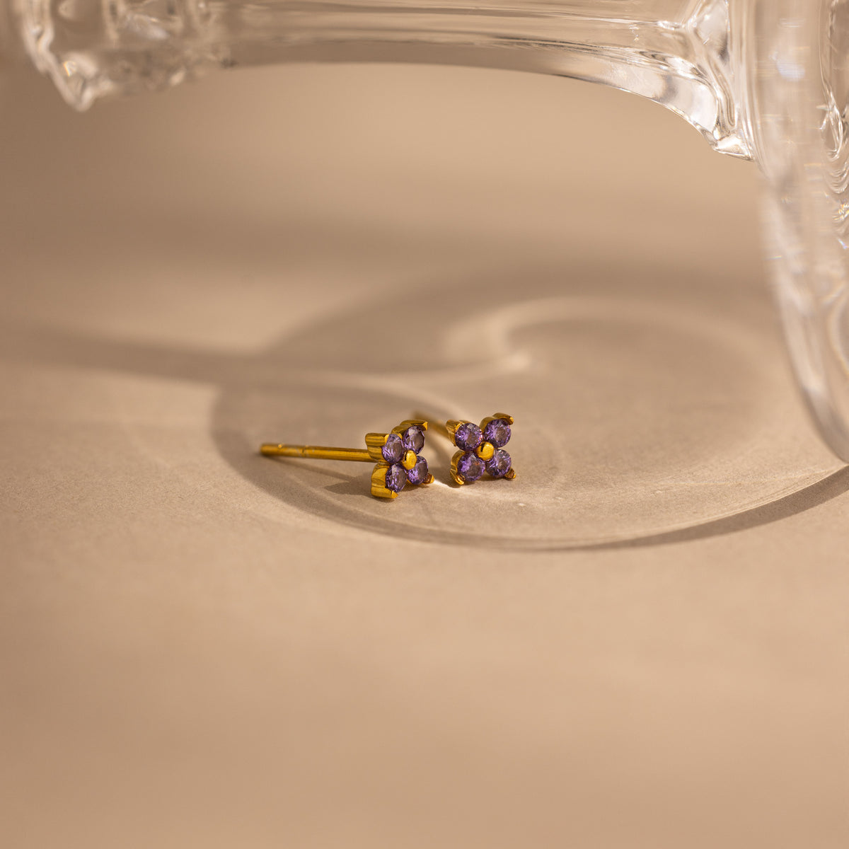A pair of small gold stud earrings with four purple gemstones arranged in a flower-like cluster, photographed on a warm beige surface with a clear glass object partially visible at the top.